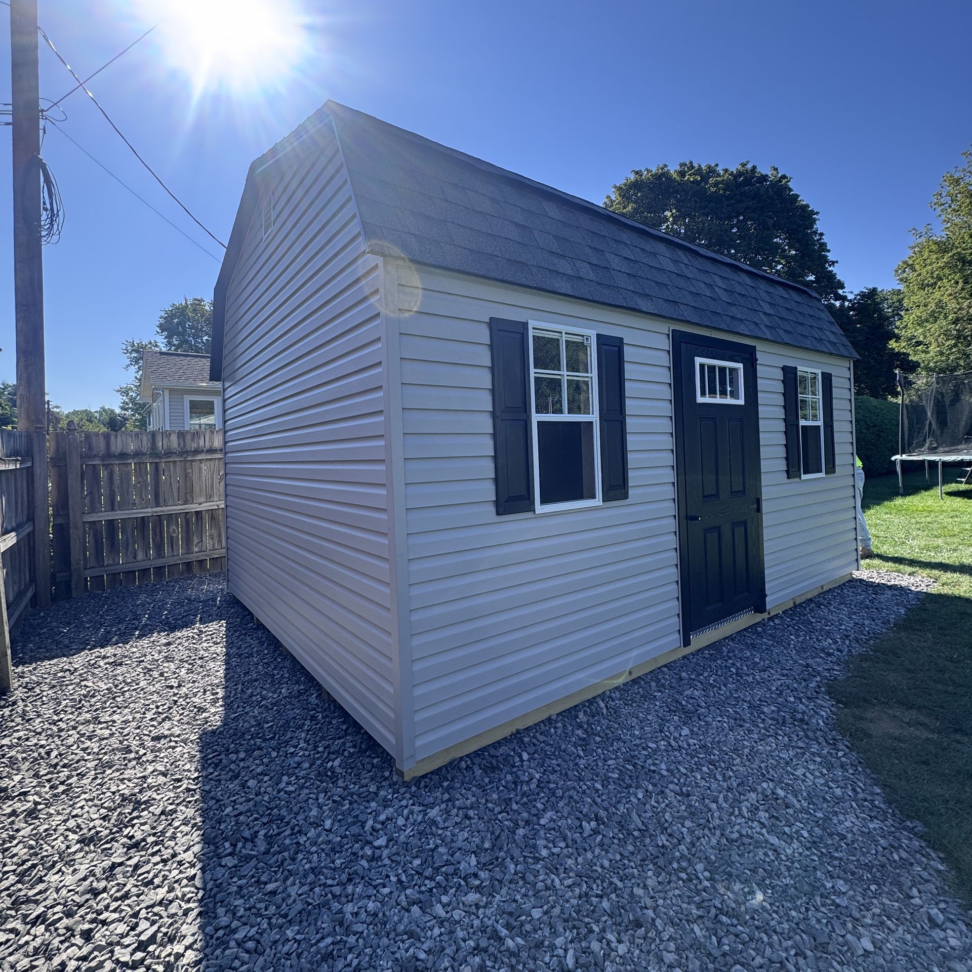 Small gray backyard shed with white trim and dark door, sitting on gravel in bright sunlight.