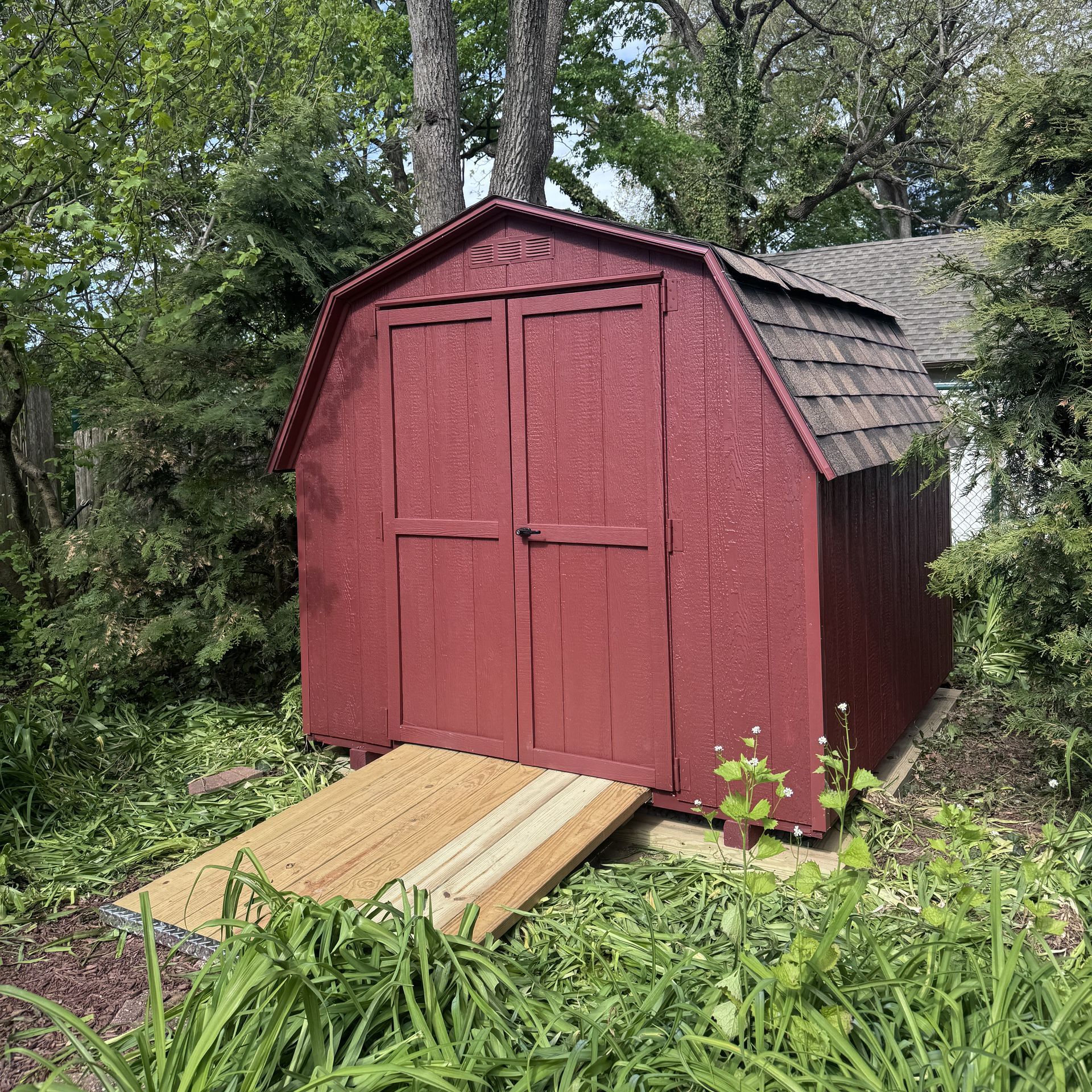 Small red wooden shed with a ramp, surrounded by greenery and trees.