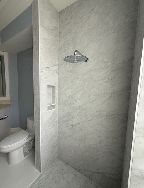 Empty tiled shower stall beside a bathroom toilet, with a wall-mounted shower head and shelf niche.