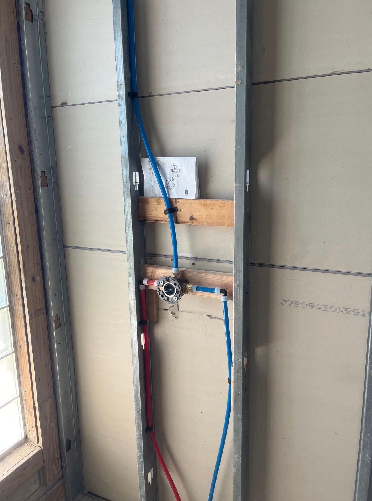 Exposed plumbing and blue wires in an unfinished wall near a window opening