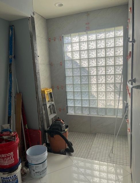 Small tiled shower room with glass block window, cleaning tools, buckets, and a vacuum cleaner.