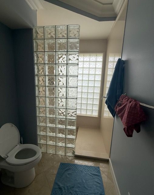 Small bathroom with toilet, glass-block shower wall, blue bath mat, and towels hanging on a rail.
