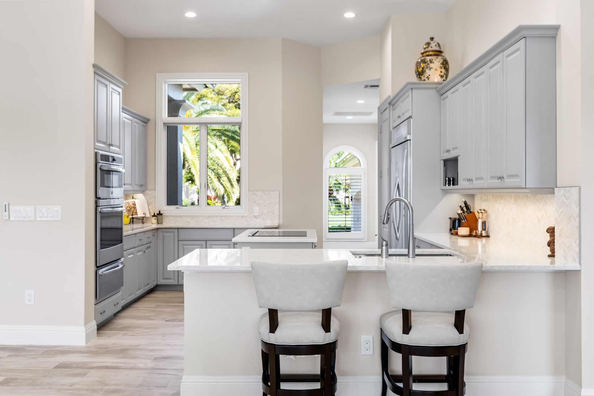 Modern kitchen with gray cabinets, white countertops, and bar stools. Bright natural light streams through a window.