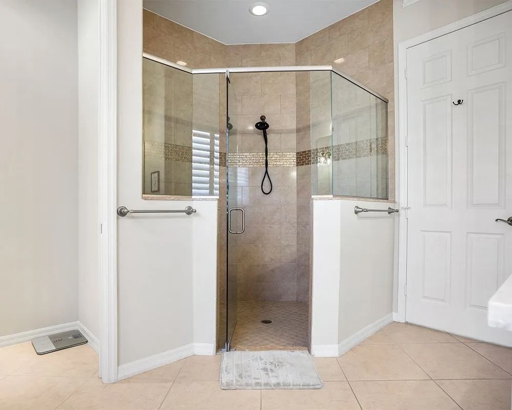 A beige-tiled, glass-enclosed shower with a gray showerhead and a white door in a bathroom.