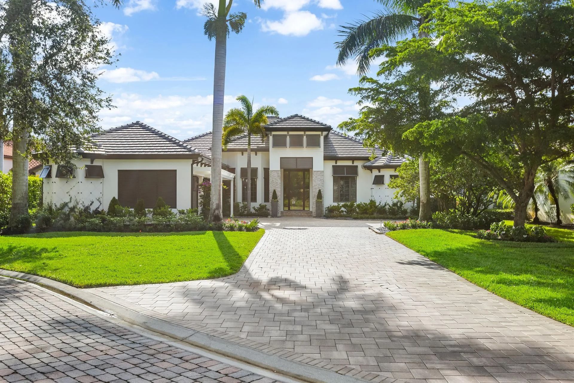 Luxurious white house with brick driveway, green lawn, palm trees, and blue sky.