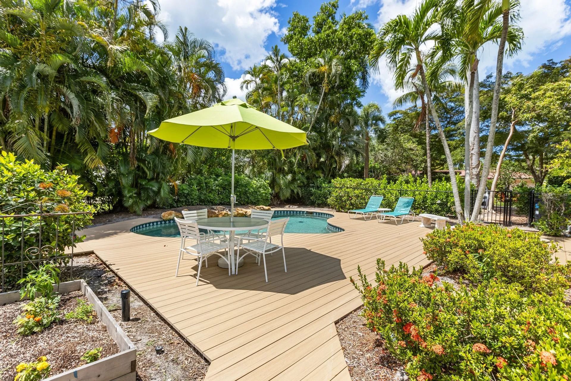 Patio with a table and umbrella by a pool, surrounded by lush green plants under a sunny sky.