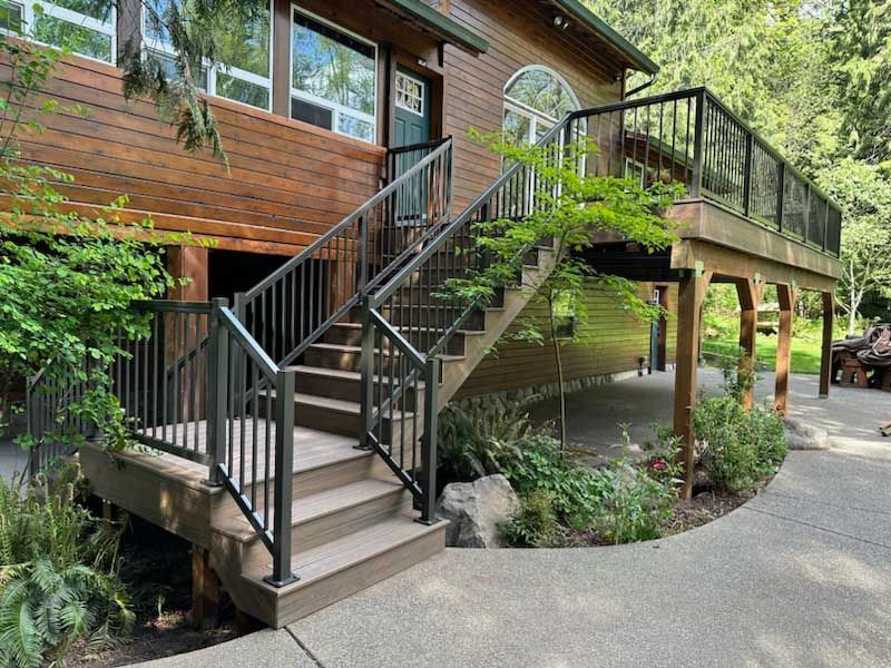 Wooden stairs and deck with black railing leading to a house in a garden setting.