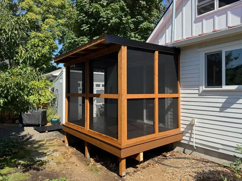 Screened-in porch addition with wood framing, attached to a white house with a black roof, in a yard.