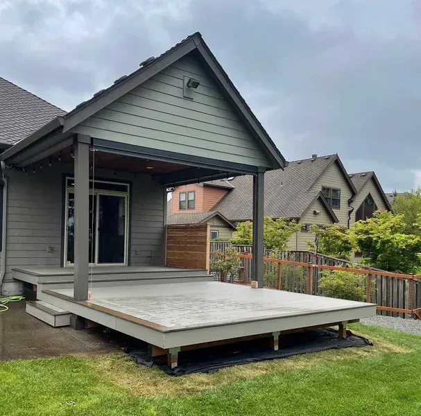 Grey deck and covered patio attached to a house with gray siding. Green grass and other houses in the background.