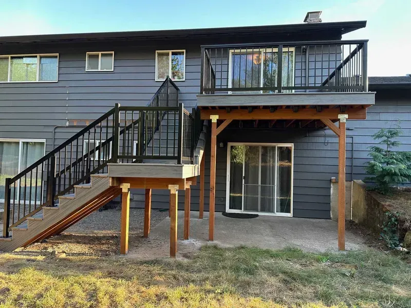 Two-story house with a wooden deck and stairs. Dark gray siding, black railing, tan support posts, and concrete patio.