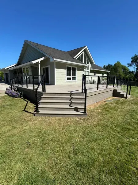 House with a large deck, gray siding, and black railing. Steps lead to the deck. Green lawn and blue sky.