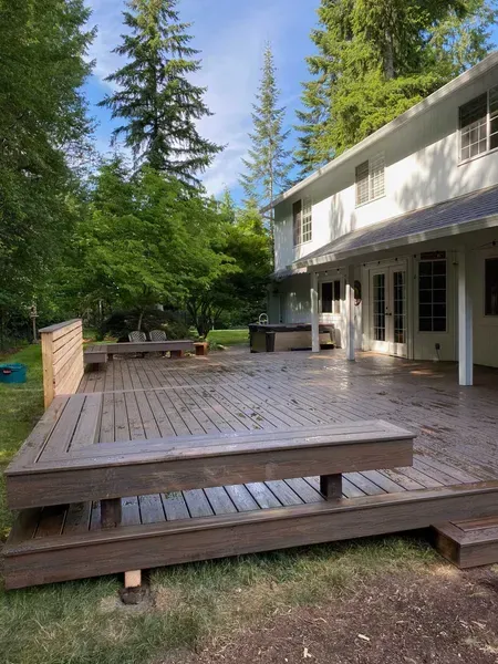 Large wooden deck with built-in bench, next to a white two-story house, surrounded by trees.