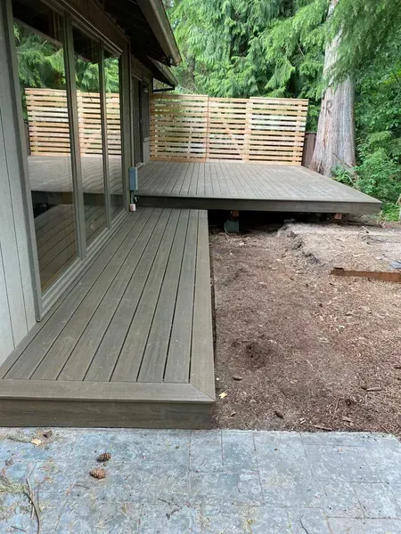 Wooden deck extension beside a house with glass windows and a natural, wooded background.