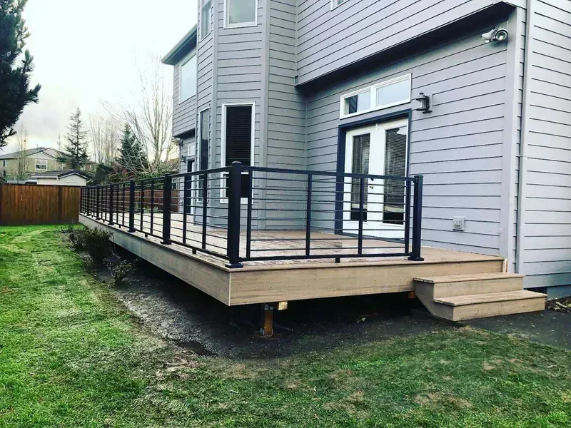 Gray house with a wooden deck, black railing, and stairs leading to a grassy yard.