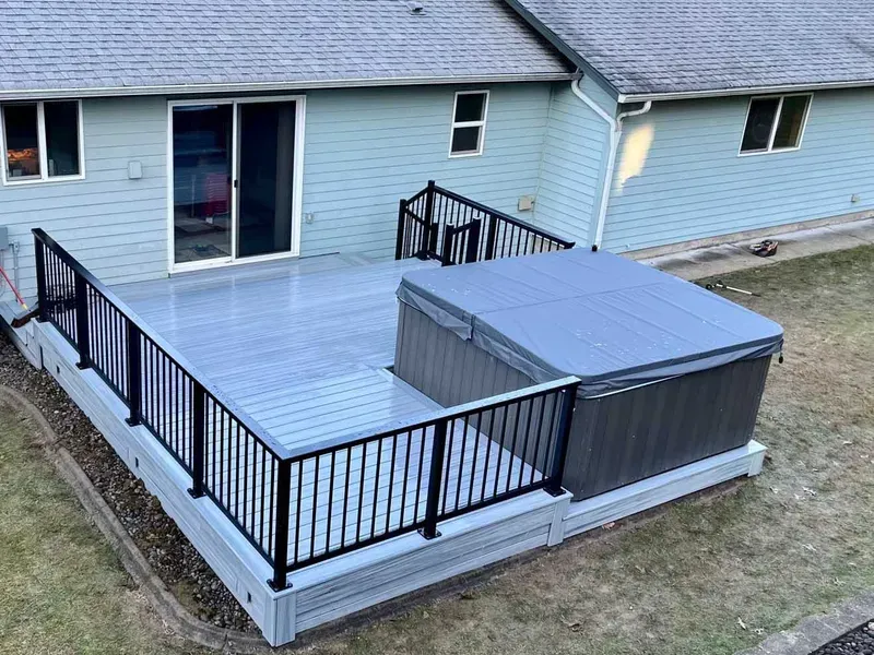 Grey composite deck with black railing and hot tub, next to a light blue house.