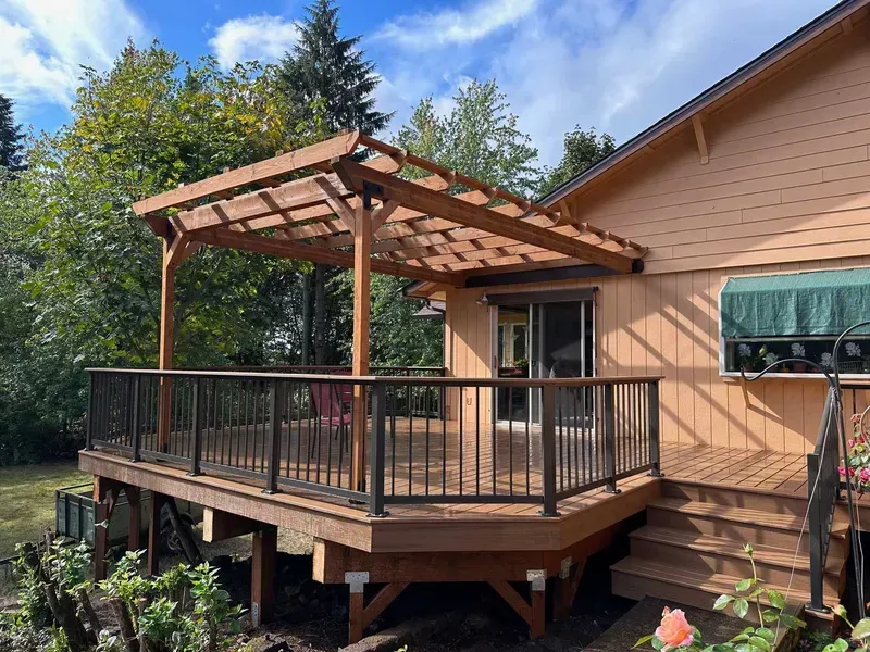 Wooden deck with pergola attached to a tan house; black railing and steps.