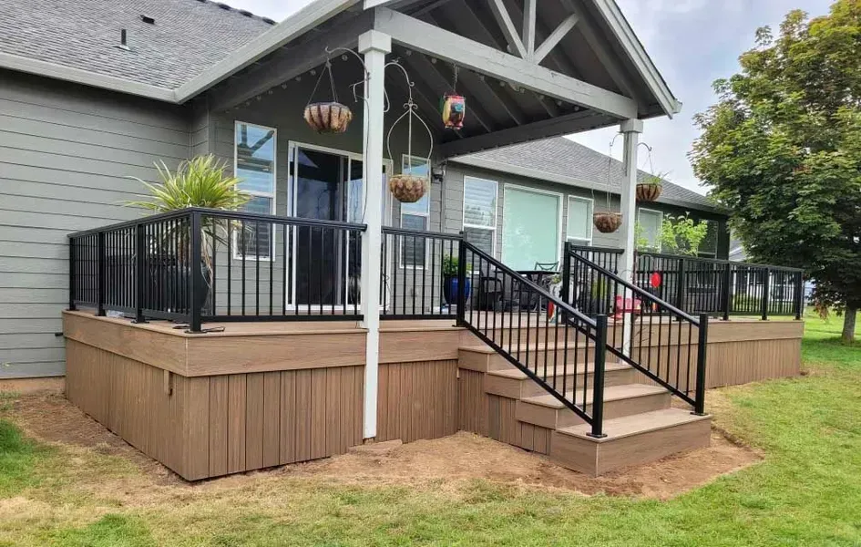 Composite deck with black railing and steps leading to a covered patio, next to a grassy lawn.