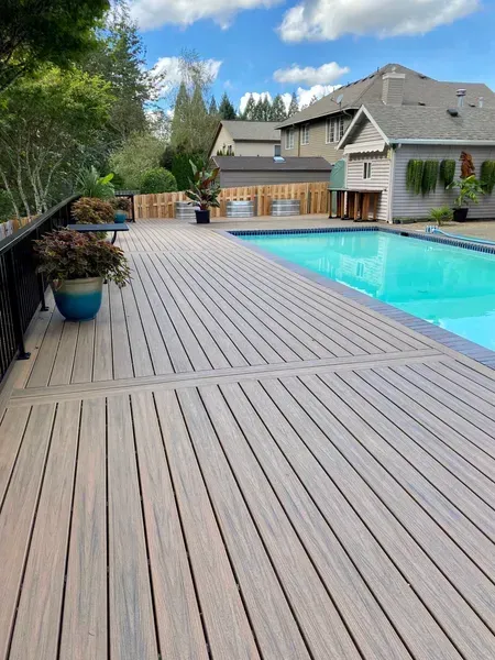 Deck and swimming pool on a sunny day. Gray deck, turquoise water, trees, and houses in the background.