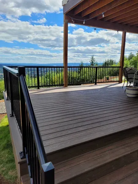 Wooden deck with black railings overlooking a scenic, wooded valley under a blue, cloudy sky.