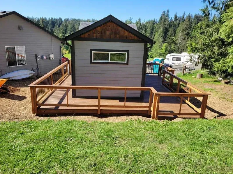 A small gray building with a wooden deck, a second house visible to the left, and a grassy yard.