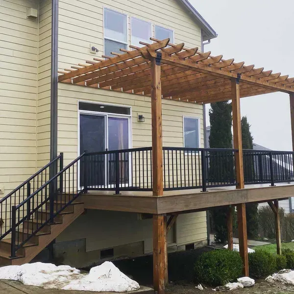 Wooden pergola over a deck attached to a light yellow house with black railings and stairs.
