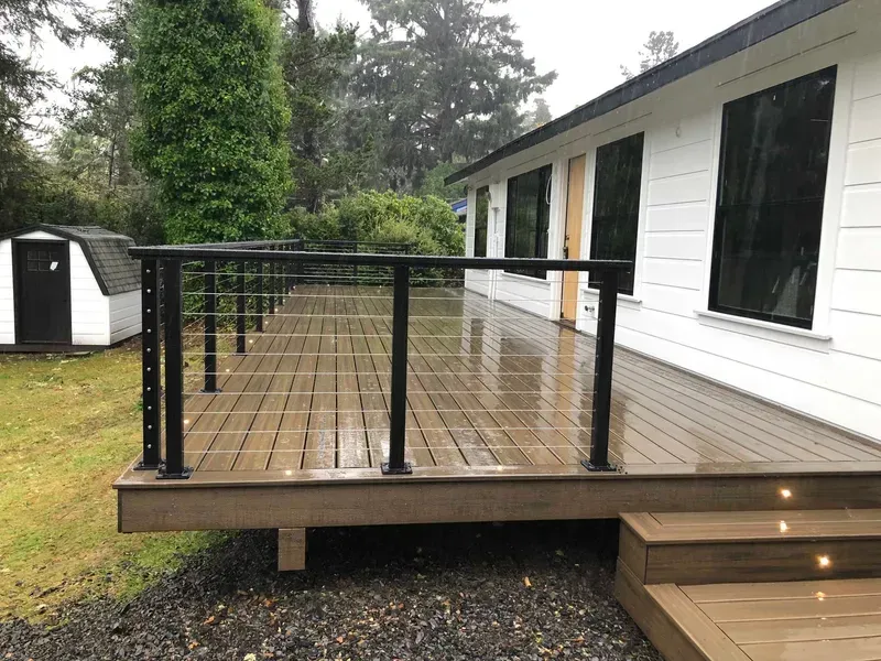 Deck with black railing and cable, attached to a white house with dark windows; rainy day.