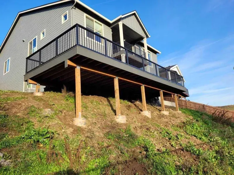 A two-story house with a wooden deck supported by posts on a grassy hillside under a clear, blue sky.