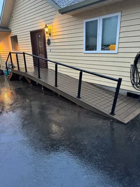 Exterior ramp with black railings leading to a doorway. Brown deck flooring, yellow siding, and wet asphalt.