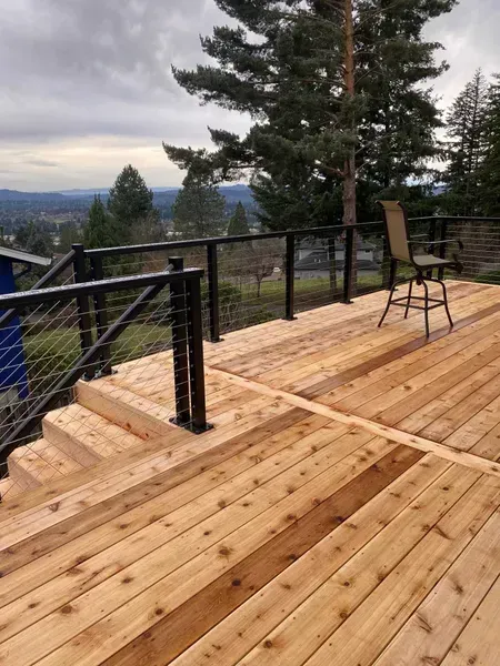 Wooden deck with steps, black railing, and cable fencing overlooking a valley under an overcast sky.