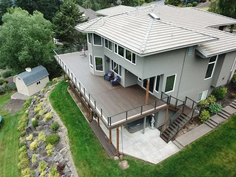 Two-story house with large deck, brown siding, and landscaping on a grassy hill.