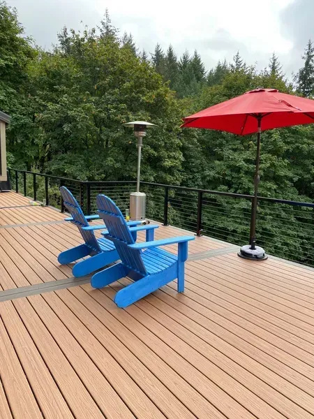 Blue Adirondack chairs on a wooden deck with a red umbrella and heater, overlooking trees.