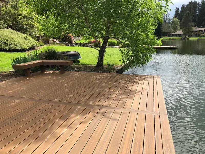 Wooden dock extending into a lake, a tree provides shade, bench, and green grass in the background.