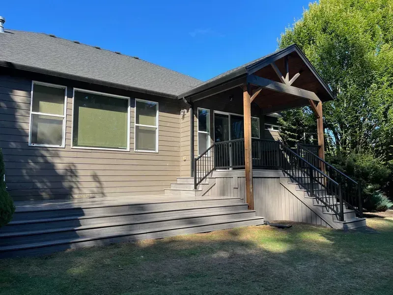 Back of a house with deck and porch, surrounded by grass and trees under a clear, blue sky.