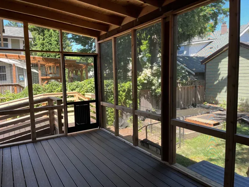 Screened-in wooden porch with dark deck and door, overlooking a backyard with greenery.