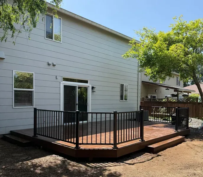 Wooden deck with black railing attached to a light gray house, sunny day.