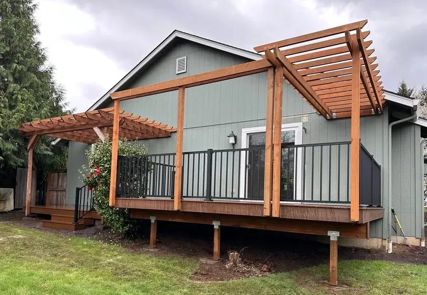 Wooden deck with pergola attached to a green house, black railing, and brown decking.