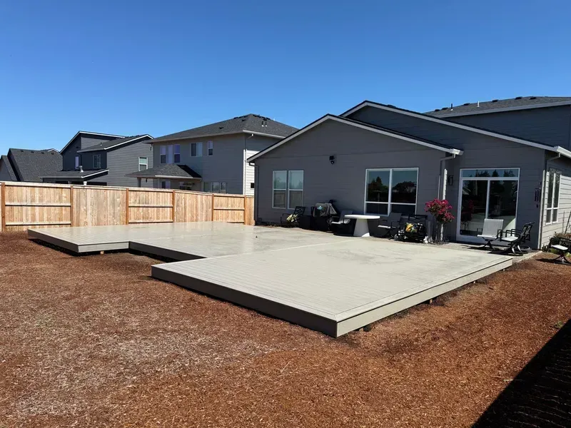 Large gray composite deck in backyard, connected to a house with a fence in the background, on a sunny day.