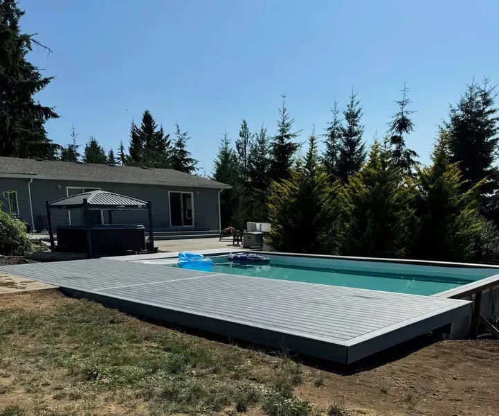 Rectangular pool with gray decking, hot tub, and house in a backyard setting under a clear blue sky.