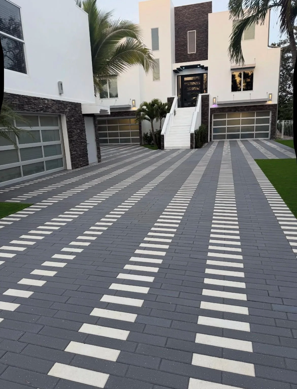 Modern residential driveway with gray pavers, white stripes, and garage doors leading to a house.