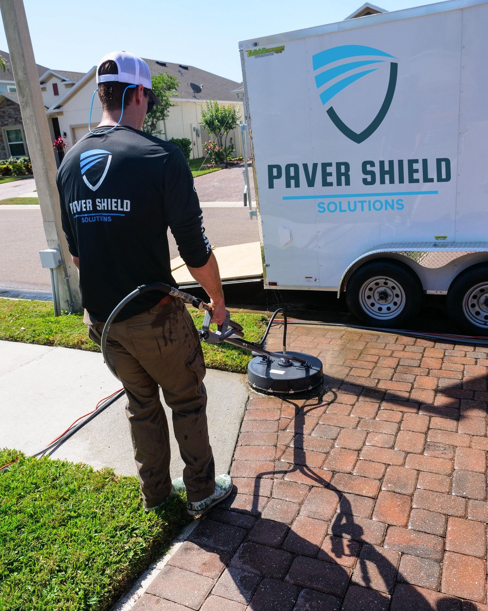 Worker power-washing brick driveway beside a Paver Shield Solutions trailer on a sunny day