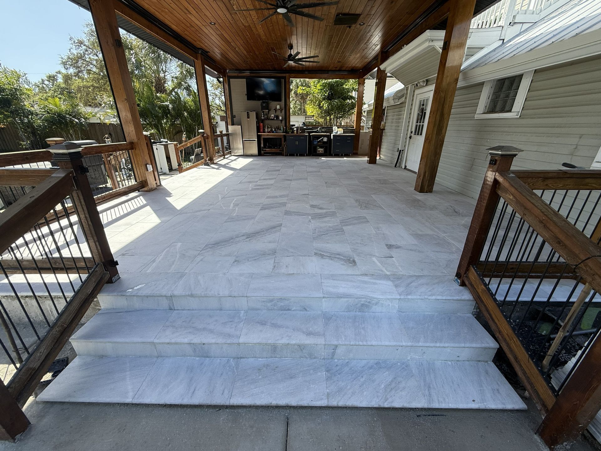Covered outdoor deck with white floor covering, wood beams, and railings leading to a patio area.