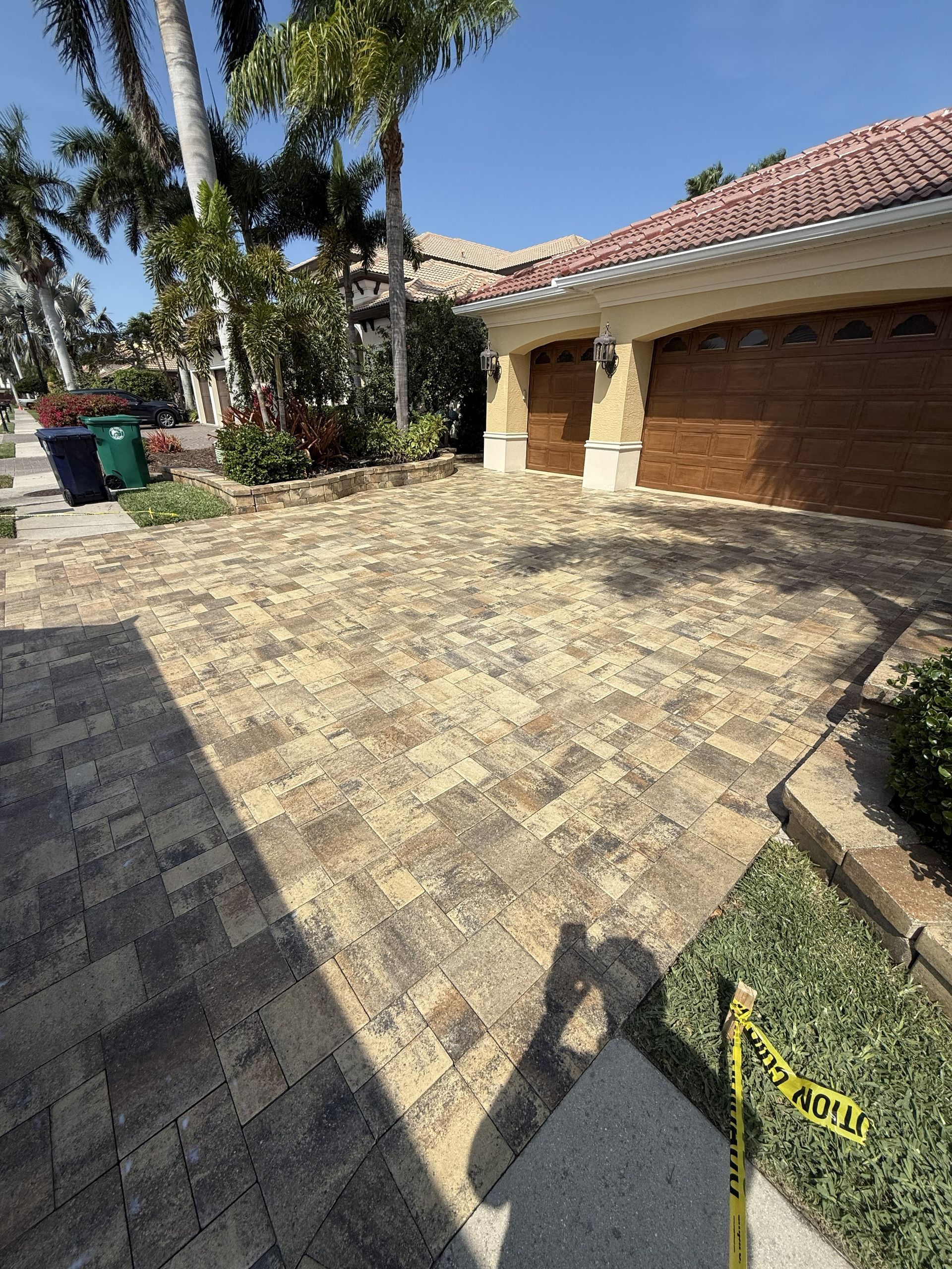 Sunlit paved driveway beside a house with a garage, palm trees, and a green trash bin