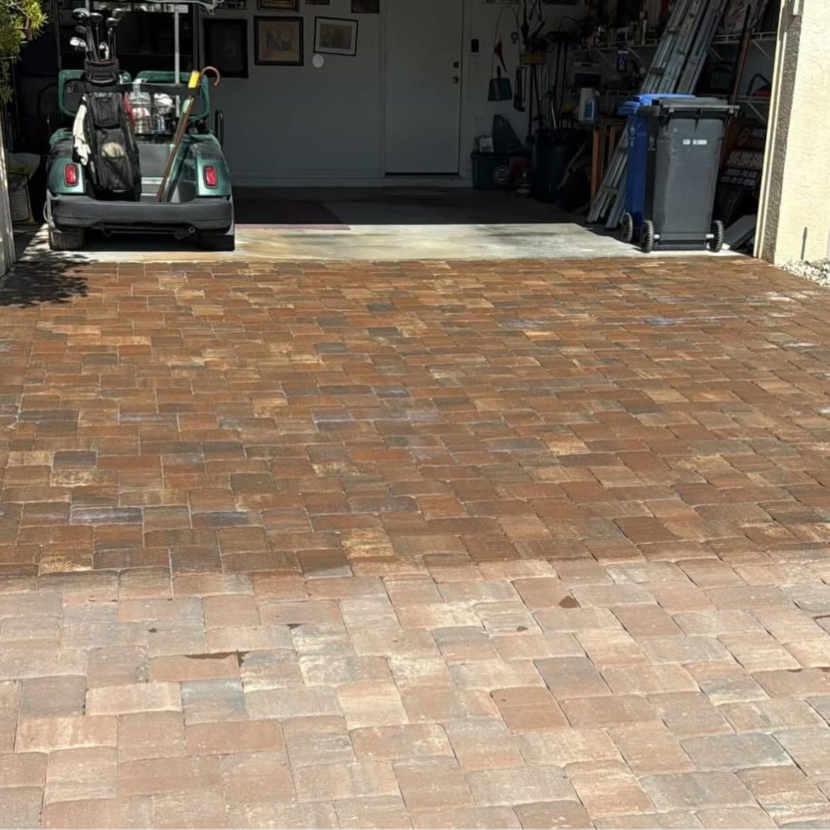 Brick driveway leading to an open garage with a lawn mower and trash bins inside