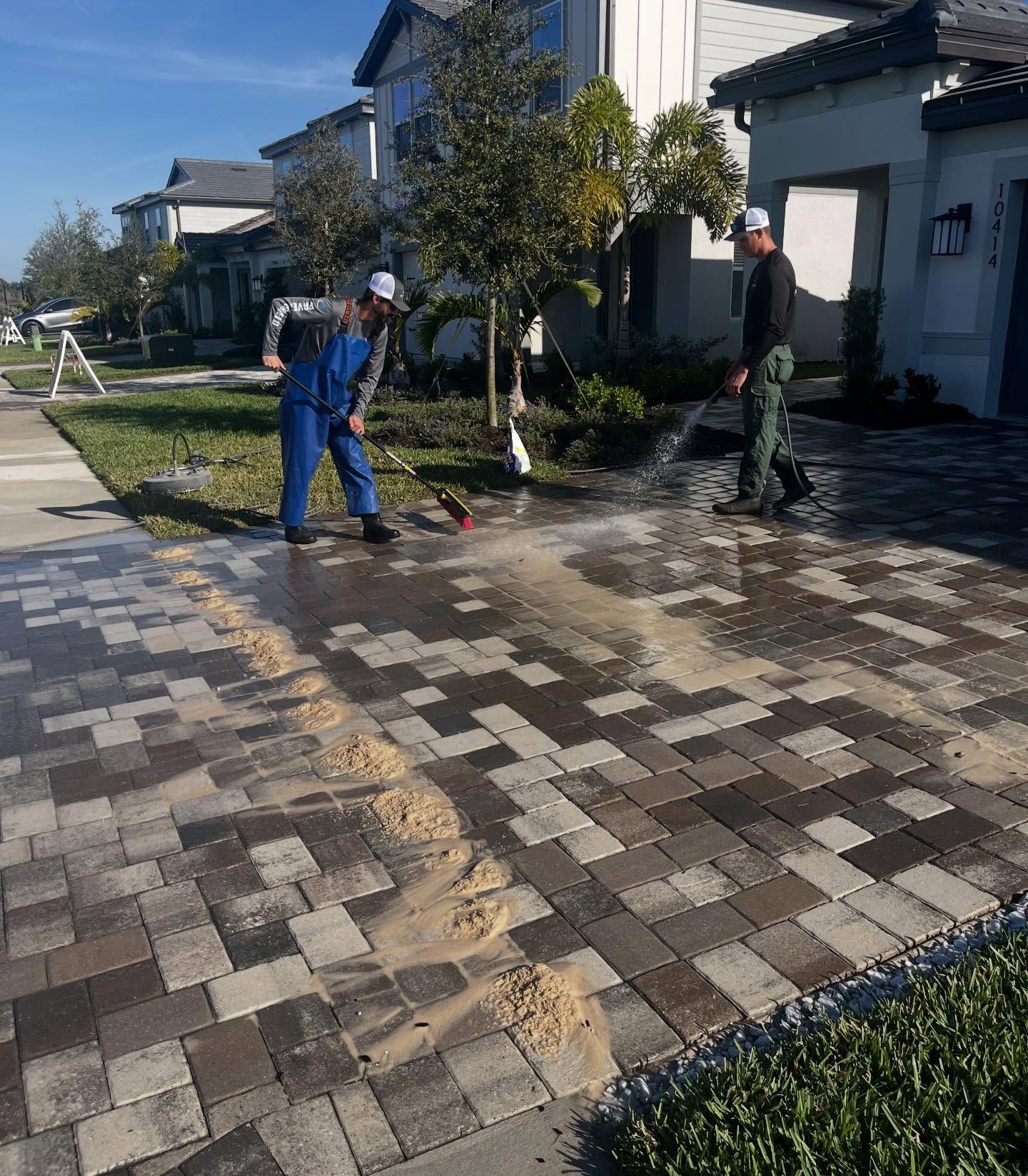 Two workers washing a patterned driveway in a sunny residential neighborhood.