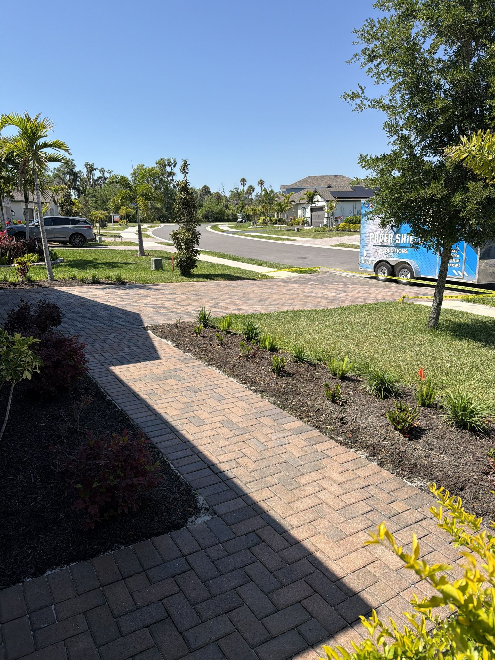 Sunny suburban sidewalk with parked cars, trees, and a blue truck near a brick path and landscaped beds