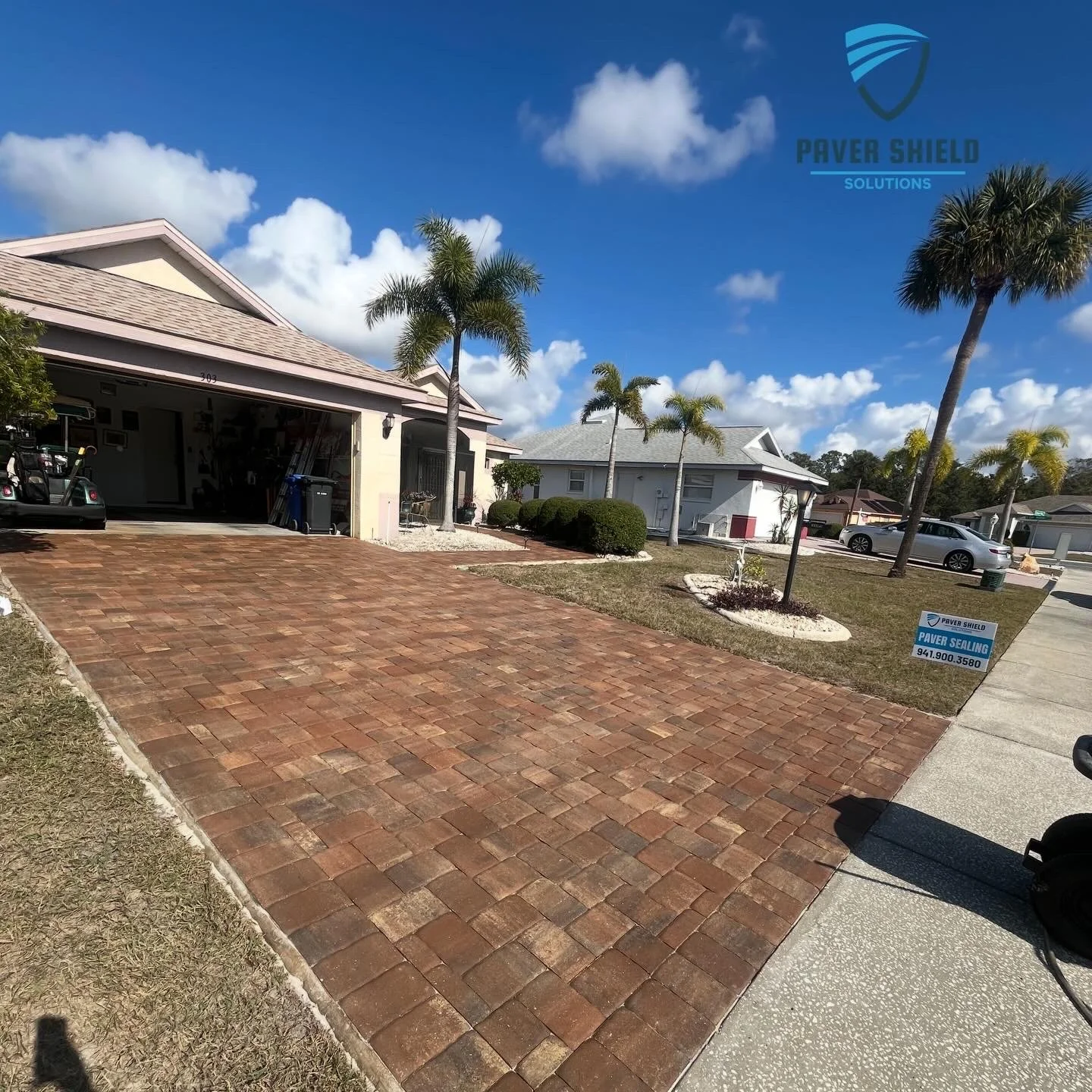 Brick driveway leading to a suburban house with palm trees under a bright blue sky