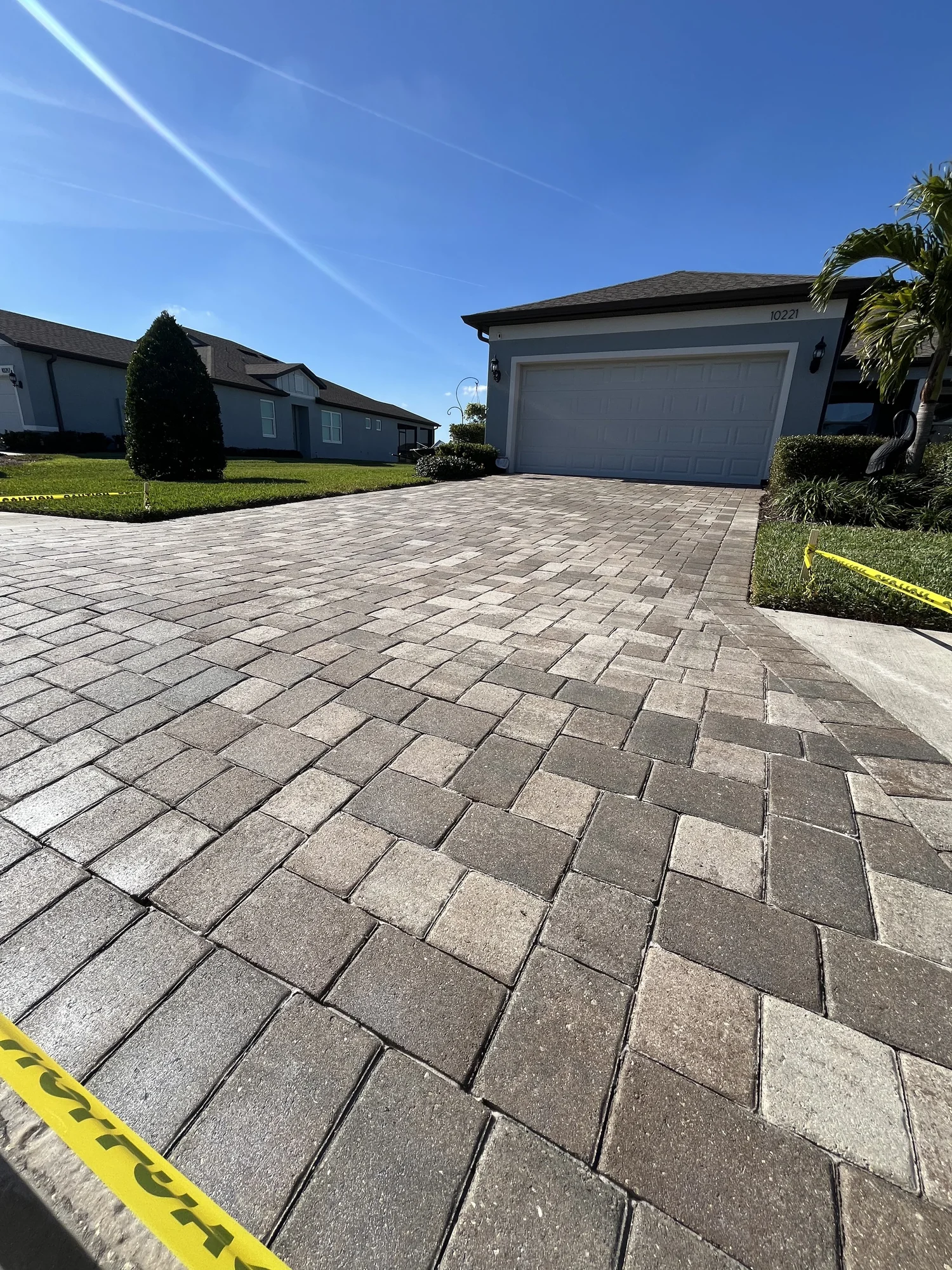 Paved driveway leading to a two-car garage under a bright blue sky, with caution tape in the foreground