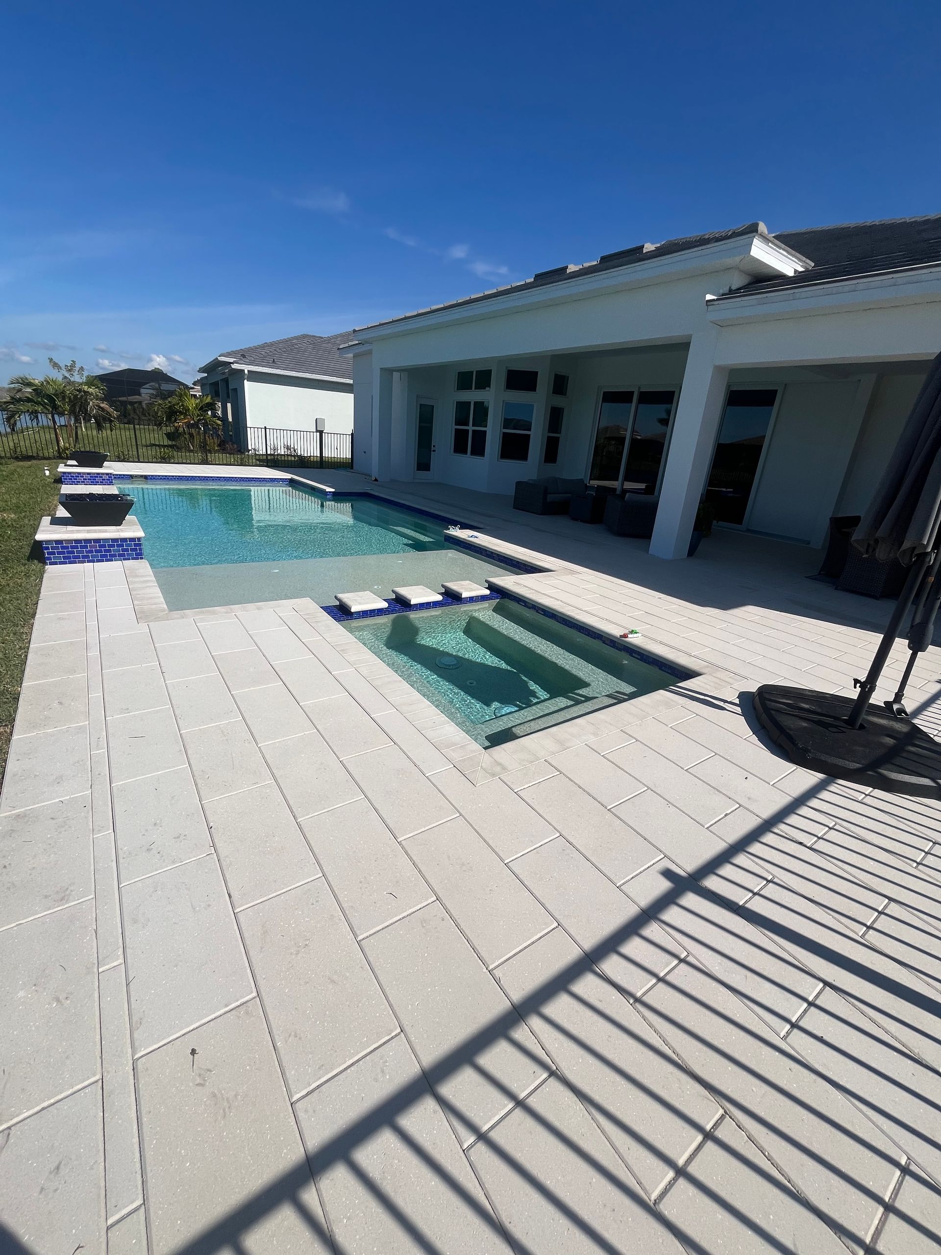 Sunny poolside patio with blue pool, white tile deck, and modern house under a clear sky