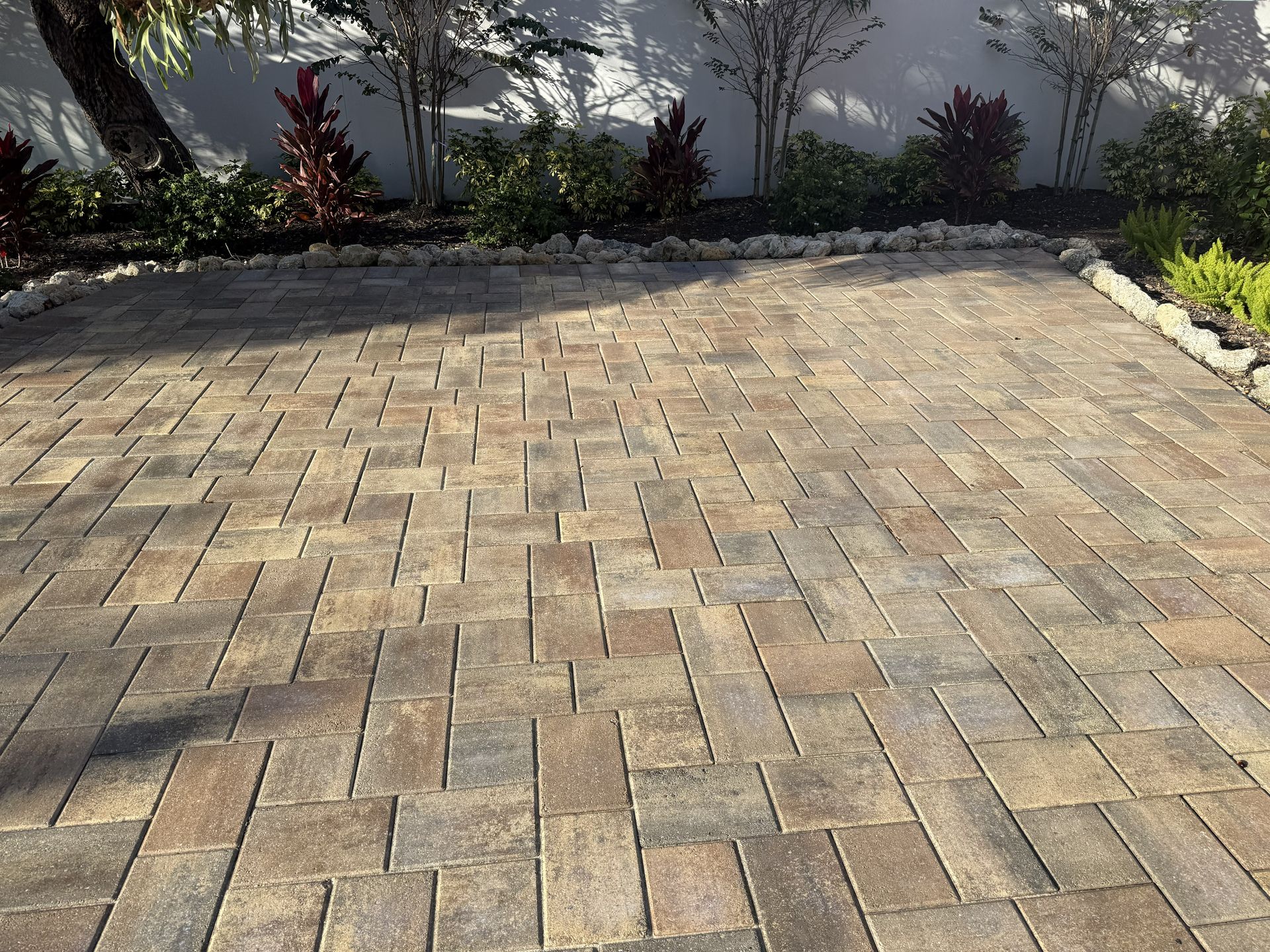 Brick patio in a garden, bordered by shrubs and a white wall in sunlight.