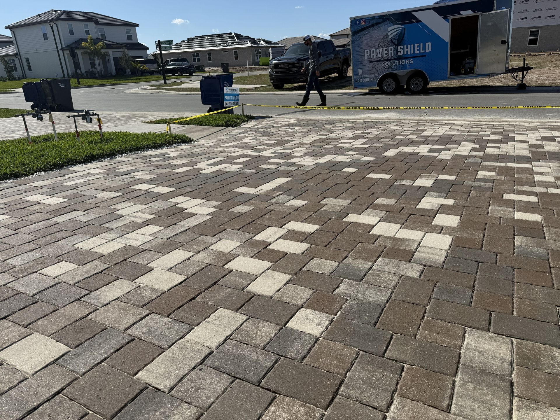 Paved courtyard with tan and gray bricks, a blue trailer, and two people walking near houses in the background